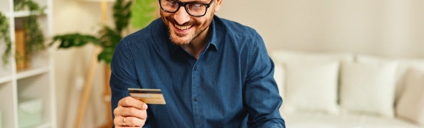 Un jeune homme heureux tient une carte de crédit dans sa main, assis à une table avec un ordinateur portable devant lui.